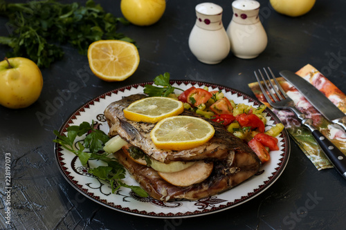 Baked fish on a plate with lemon, salad and greens. On the table are parsley, apples, lemon, knife, fork, salt and pepper