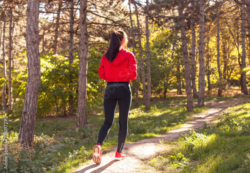 Fototapeta Naklejka Na Ścianę i Meble -  A beautiful, young girl running through the woods during sunset. Athletic woman in sportswear running along a forest trail. Portrait, full length with the forest in the background. View from the back
