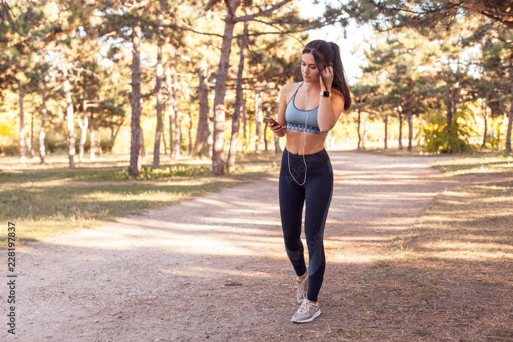 A beautiful, young girl running through the woods during sunset. The girl in the headphones plays sports and listens to music on the phone. Fitness sport woman in fashion sportswear. Outdoor sports