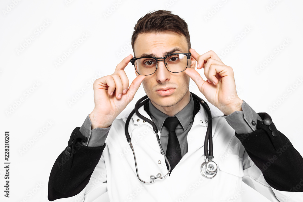 A focused doctor in a white robe and a stethoscope around his neck looks at his glasses, trying to see something, looking at the camera isolated on a white background