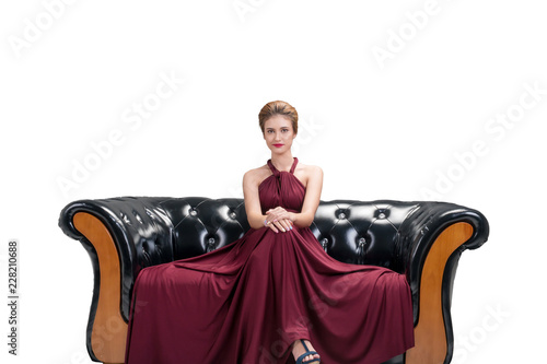 Attractive young woman wear red dress fashion posing and sitting on black sofa looking at camera on isolated white background. Beauty and luxurious concept