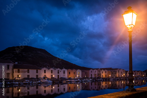 Night city view of Bosa harbor, Sardinia, Italy with lantern switched on. Colorful houses and blue sky reflecting in the water. 