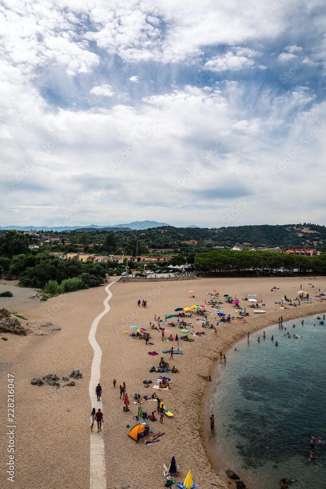 People sunbathing on the beach of Torre di Bari, Sardinia, Italy. Beautiful blue sky with clouds and mountains in the background and a path visible.