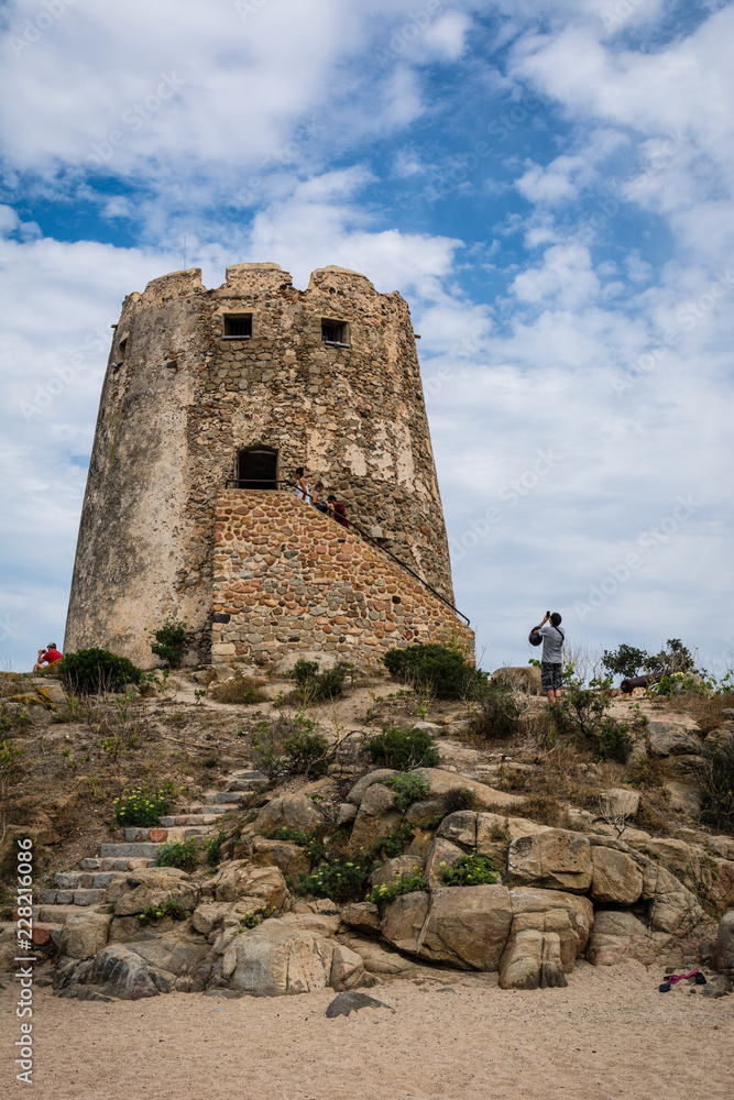 Old watchtower on the beach of Torre di Bari, Sardinia, Italy. Beautiful blue sky with clouds in the background, a path visible and some unrecognizable people.