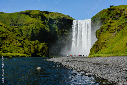 Famous Skogafoss Waterfall, Iceland with colourful tourists visible by water, sunny summer day, blue sky