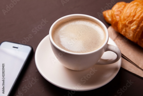 Fototapeta Naklejka Na Ścianę i Meble -  Coffee break, morning coffee. Cup of coffee with foam and croissant with smartphone.