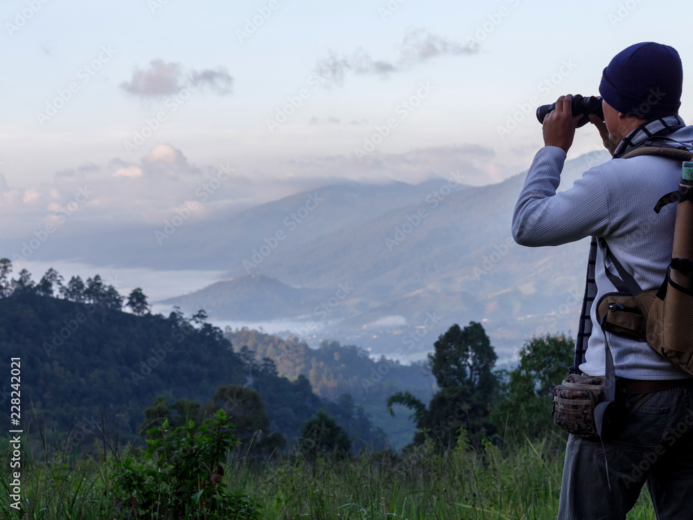 Young man with backpack and holding a binoculars looking on top of mountain Young man with backpack and holding a binoculars looking on top of mountain