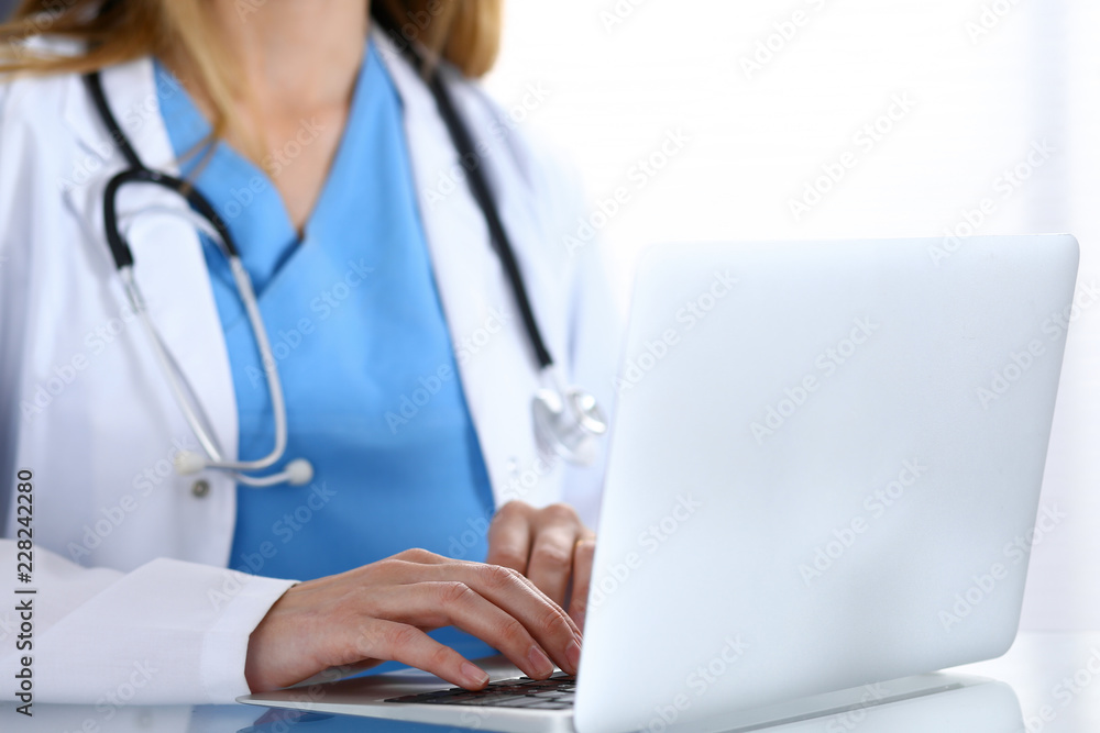 Doctor typing on laptop computer while sitting at the glass desk in hospital office. Physician at work. Medicine and healthcare concept Doctor typing on laptop computer while sitting at the glass desk in hospital office. Physician at work. Medicine and healthcare concept