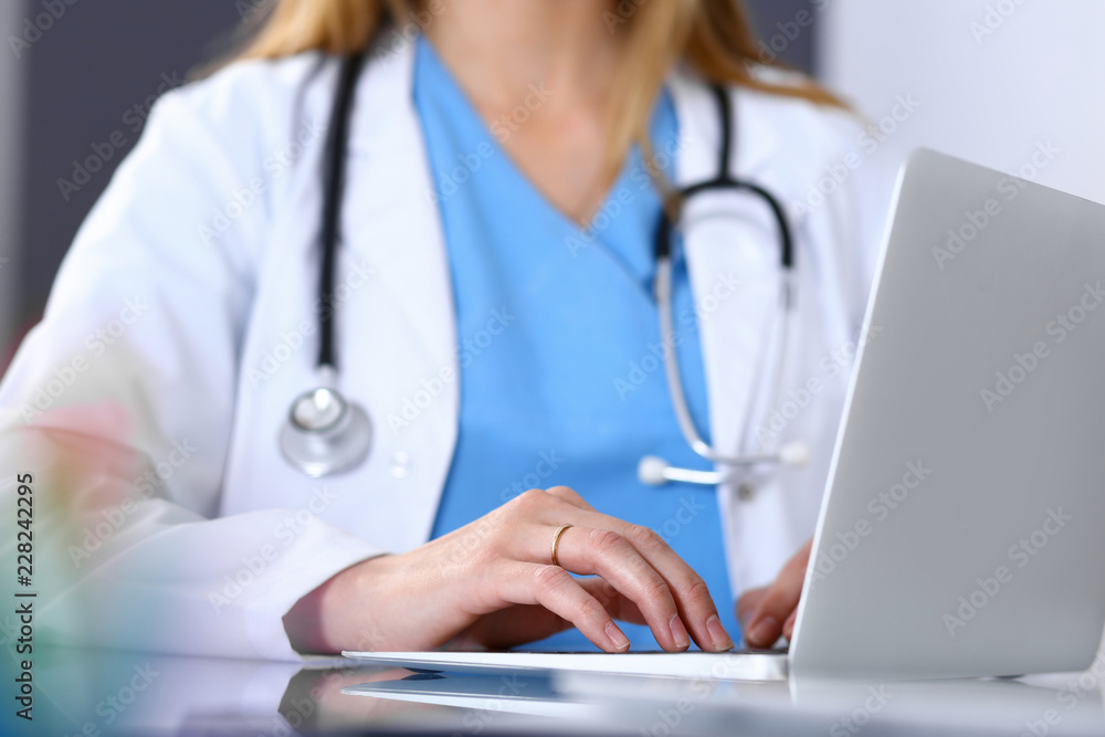 Doctor typing on laptop computer while sitting at the glass desk in hospital office. Physician at work. Medicine and healthcare concept Doctor typing on laptop computer while sitting at the glass desk in hospital office. Physician at work. Medicine and healthcare concept