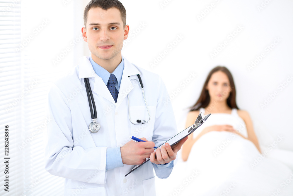Medicine doctor standing and smiling on the background with patient in the bed Medicine doctor standing and smiling on the background with patient in the bed