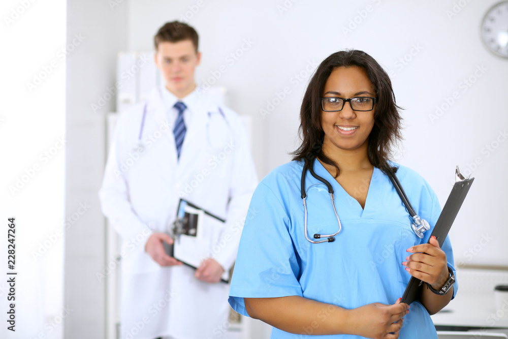Female african american medical doctor with colleagues in background at hospital. Medicine and health care concept Female african american medical doctor with colleagues in background at hospital. Medicine and health care concept