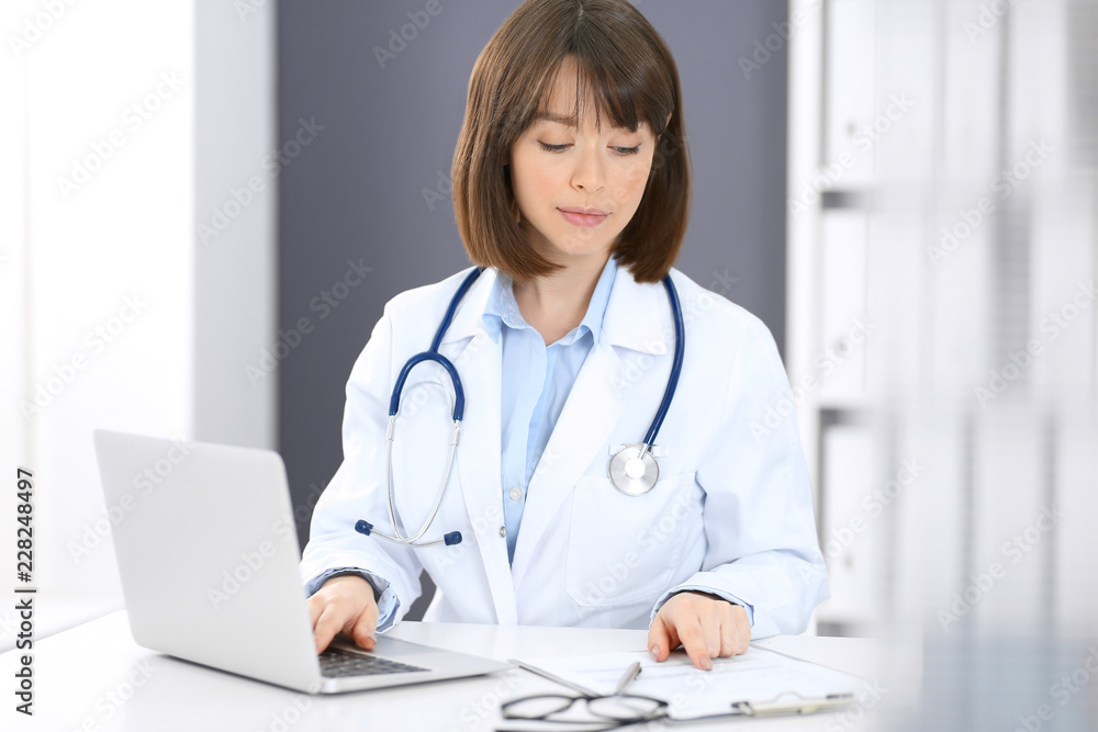 Doctor typing on laptop computer while sitting at the white table in hospital office. Physician at work. Medicine and healthcare concept Doctor typing on laptop computer while sitting at the white table in hospital office. Physician at work. Medicine and healthcare concept