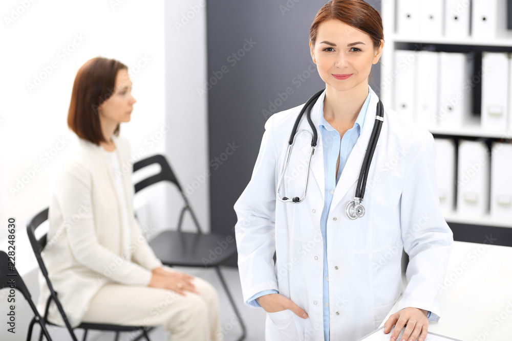 Doctor woman at work. Portrait of female physician cheerful smiling while standing near reception desk at clinic or emergency hospital. Patient woman sitting at the background. Medicine concept Doctor woman at work. Portrait of female physician cheerful smiling while standing near reception desk at clinic or emergency hospital. Patient woman sitting at the background. Medicine concept