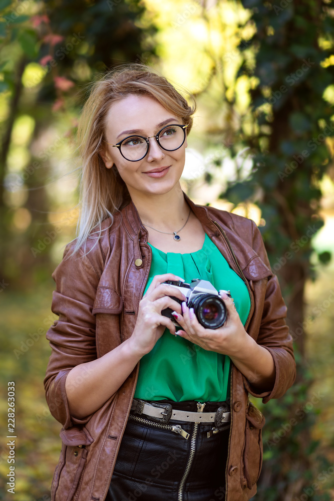 Outdoor portrait of beautiful young woman having fun in park with retro camera. Photographer with hipster style glasses taking pictures
