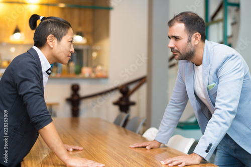 Papier peint Tense Latin and Asian colleagues standing at desk looking at each other and keeping hands on desk surface