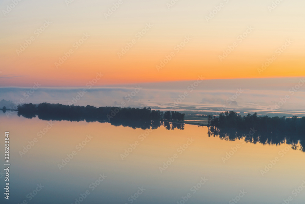 Broad mystical river flows along diagonal shore with silhouette of trees and thick fog. Gold glow in predawn sky. Calm morning atmospheric landscape of majestic nature in warm tones.