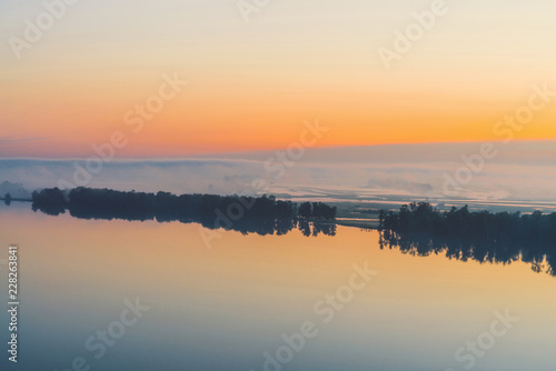 Broad mystical river flows along diagonal shore with silhouette of trees and thick fog. Gold glow in predawn sky. Calm morning atmospheric landscape of majestic nature in warm tones. © Daniil