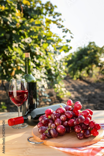 Fresh grape with bottles and glass of red wine on wooden table in vineyard