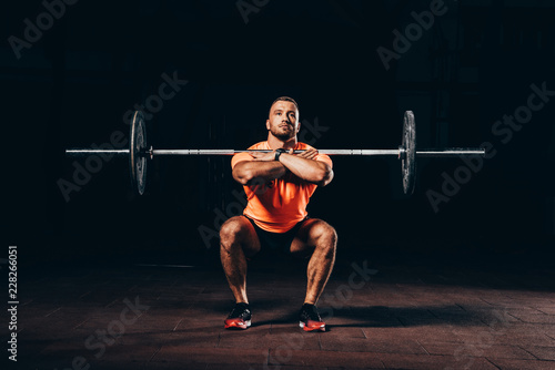 handsome muscular man doing squats with barbell in dark gym