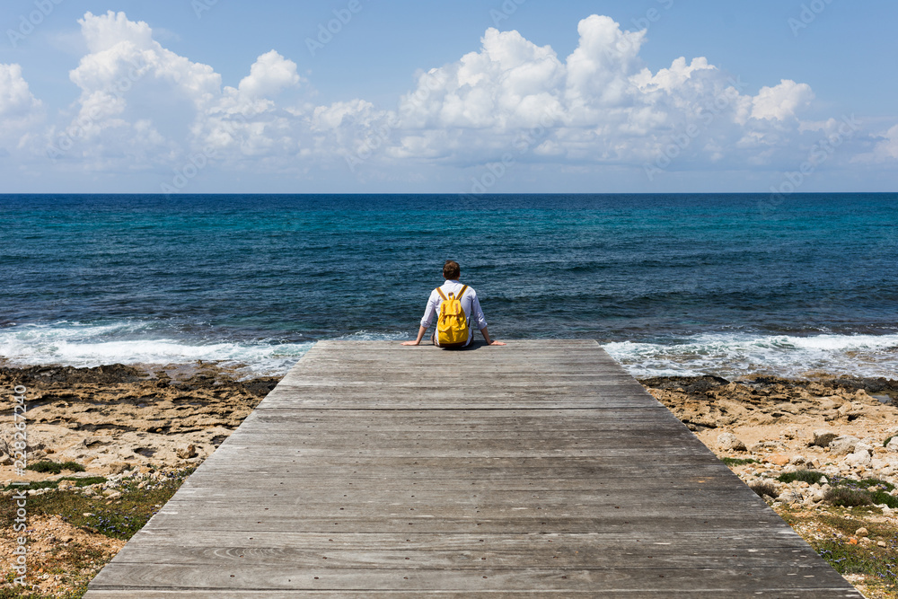 Tourist traveler guy stands on a wooden pier in solitude and looks at the blue sea, ocean. Calm and peace. Yellow backpack. The coast and a sandy beach with stones. Travel and vacation with a hike.