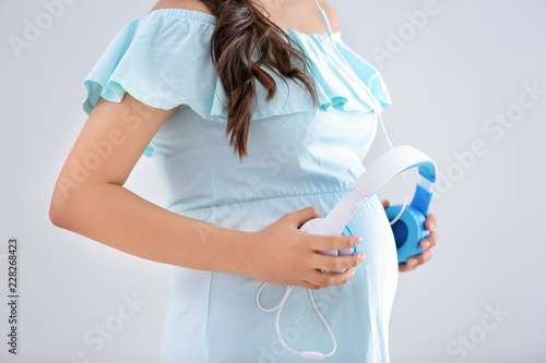 Beautiful pregnant woman putting headphones on her belly against light background