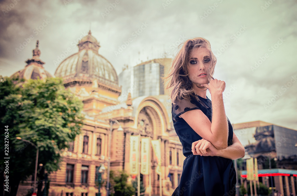 Fototapeta premium Beautiful blonde girl with big blue eyes and black dress and messed up hair, looking pensive, in the city