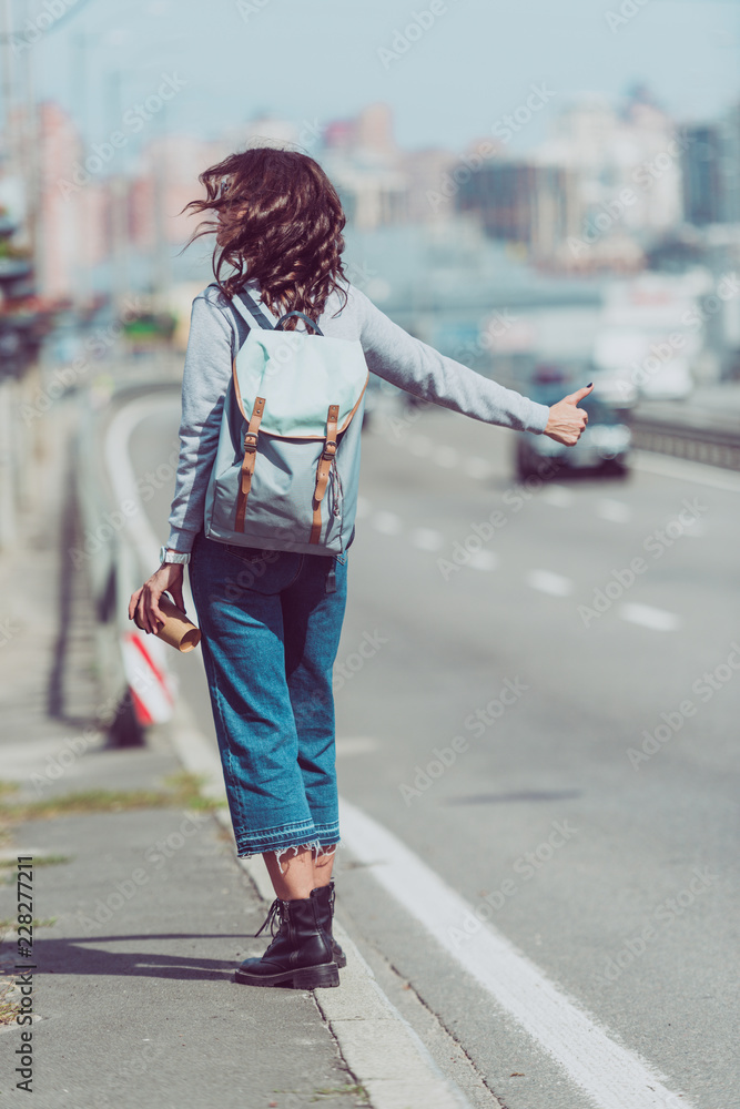 rear view of woman with backpack and coffee to go hitchhiking at road