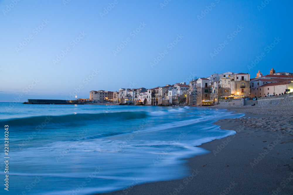 Panoramic view of Cefalù after sunset, with lights and small waves breaking on the beach