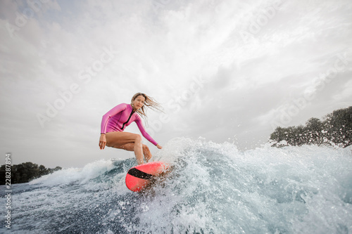 Blonde woman wakesurfer riding down the blue splashing wave against sky