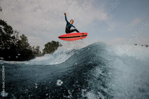 Happy emotional boy dressed in black and blue swimsuit wakesurfing jumping up on orange board