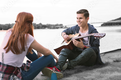 Cheerful boy. Handsome boy with guitar feeling cheerful while sitting near river with his appealing red-haired friend © Viacheslav Yakobchuk