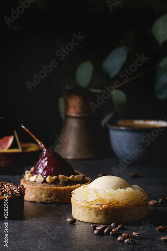 Variety of sweet tartlets with chocolate, caramel, pears, figs with cup of coffee and coffee beans around on black texture table.
