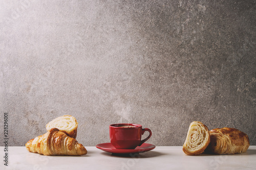 Fresh baked whole and sliced croissant with red cup of coffee espresso on white marble table with grey wall at background. Caffe breakfast