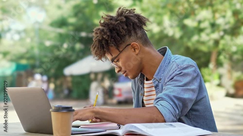 Smiling african student in eyeglasses using laptop computer and writing somthing while sitting by the table in cafe outdoors