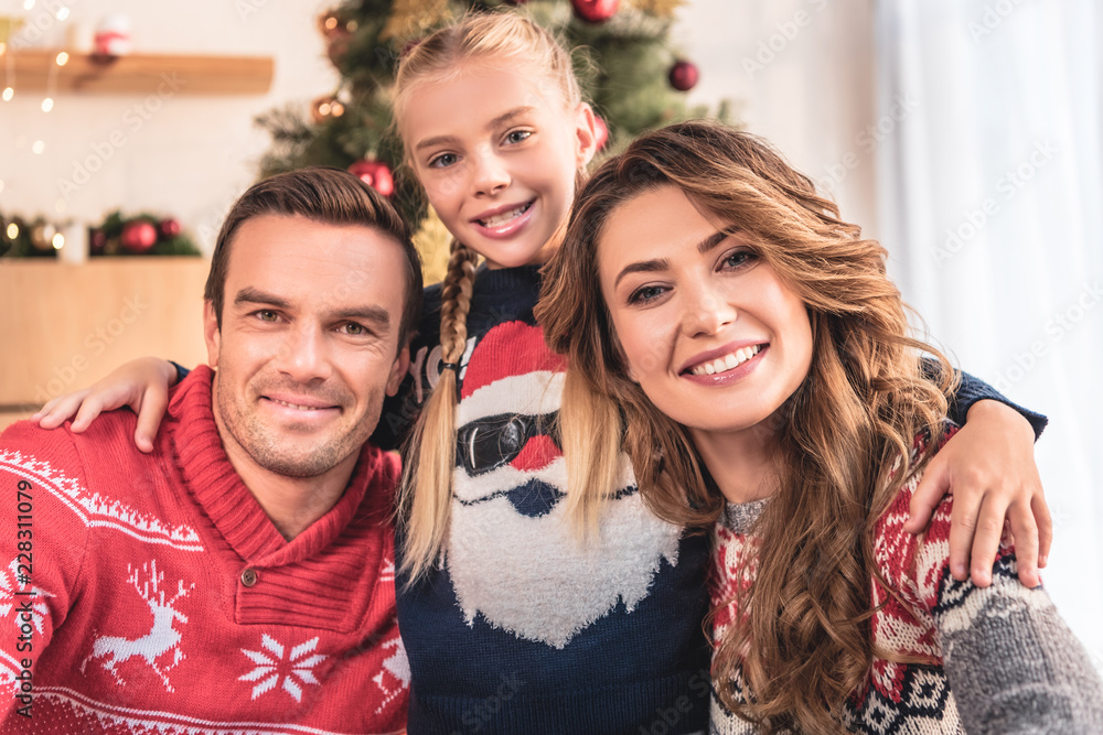 Obraz premium smiling parents and daughter in winter sweaters hugging and looking at camera at home