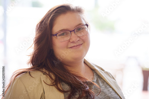 Photography close up portrait of beautiful plus size young adult woman, with brown hair and a beige blazer, while looking into the camera with her prescription glasses on