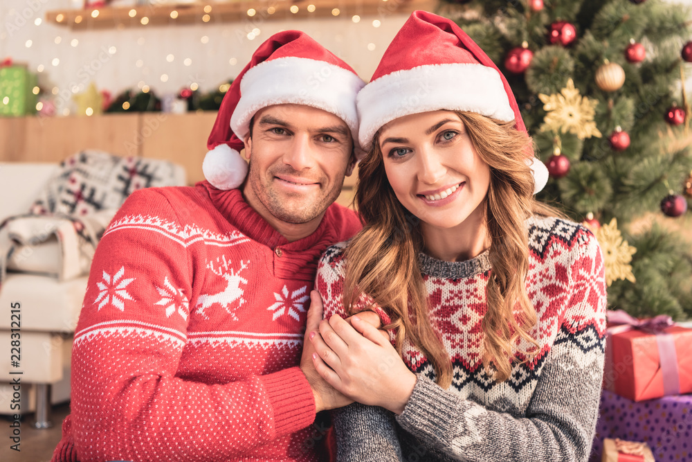 smiling husband and wife in santa hats holding hands near cristmas tree at home, looking at camera smiling husband and wife in santa hats holding hands near cristmas tree at home, looking at camera