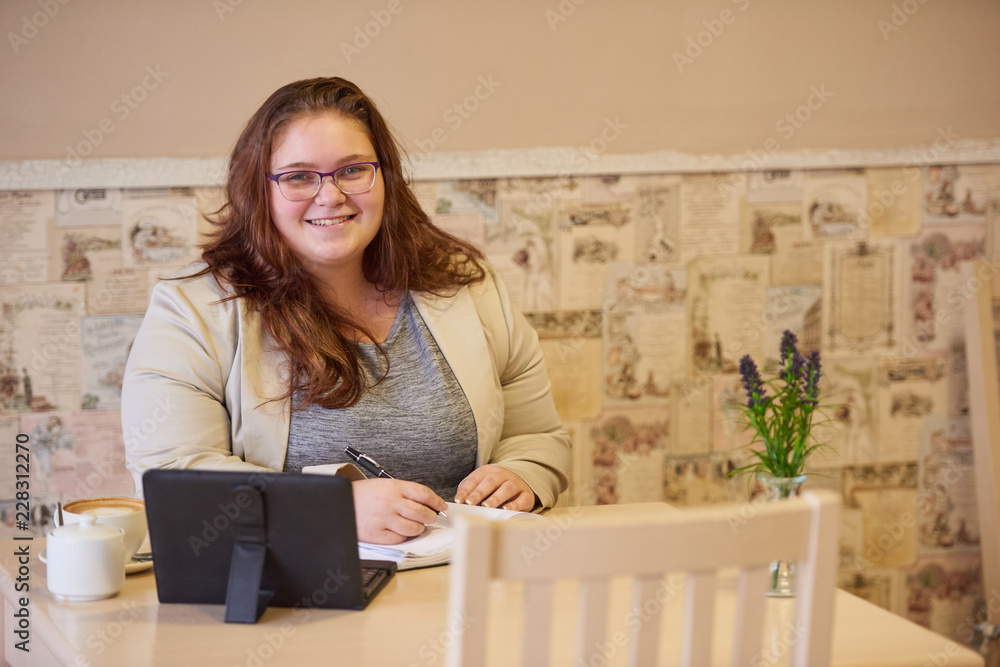 Plus size caucasian businesswoman with brown hair hanging loose, while smiling at camera in a coffee shop decorated with warm tones creating a cosy atmosphere. Plus size caucasian businesswoman with brown hair hanging loose, while smiling at camera in a coffee shop decorated with warm tones creating a cosy atmosphere.