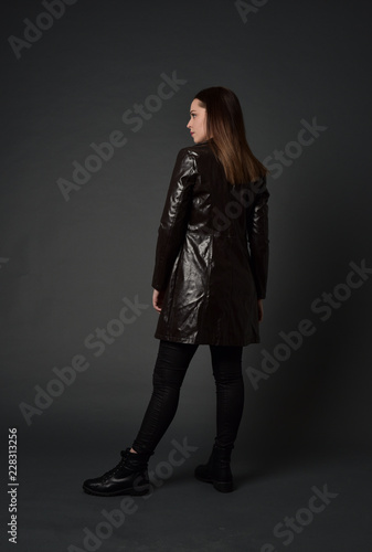 full length portrait of brunette woman wearing long leather coat. standing pose with back to the camera, on grey studio background.