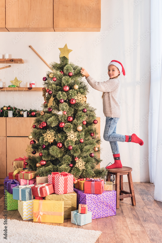 smiling kid in santa hat standing on stool and decorating christmas tree at home with presents