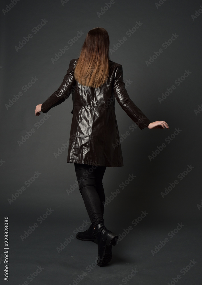 full length portrait of brunette woman wearing long leather coat. standing pose with back to the camera, on grey studio background. full length portrait of brunette woman wearing long leather coat. standing pose with back to the camera, on grey studio background.