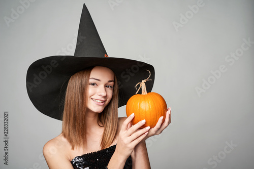 Tablou canvas Closeup of smiling Halloween witch holding orange pumpkin, isolated on grey back