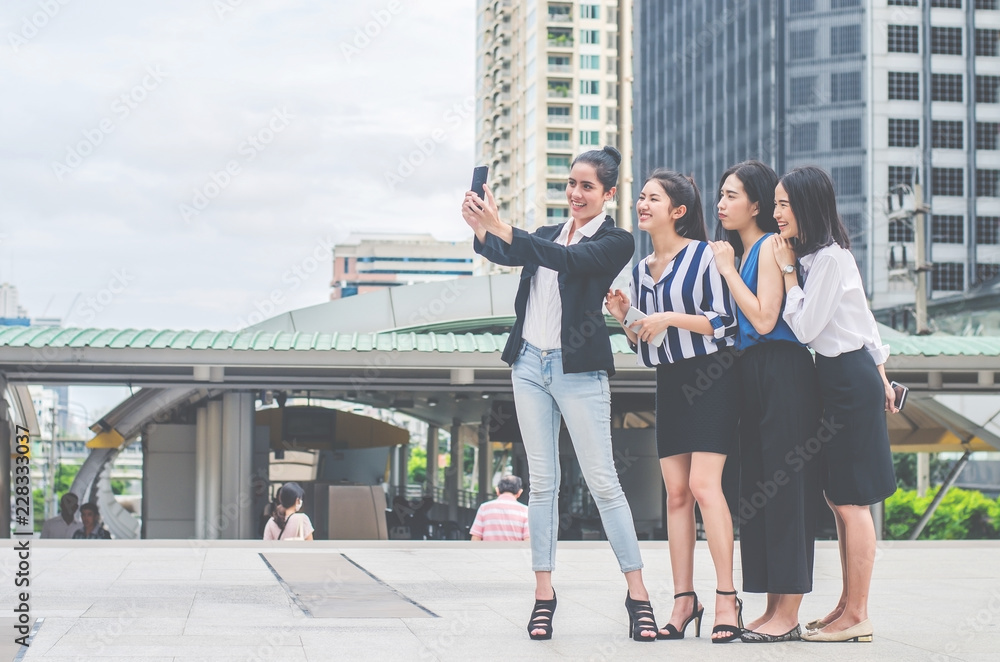 Obraz premium Four happy Asian businesswoman colleague taking selfie with smartphone in city,looking at camera, happy healthy diverse fit people relaxing after working out together in office.