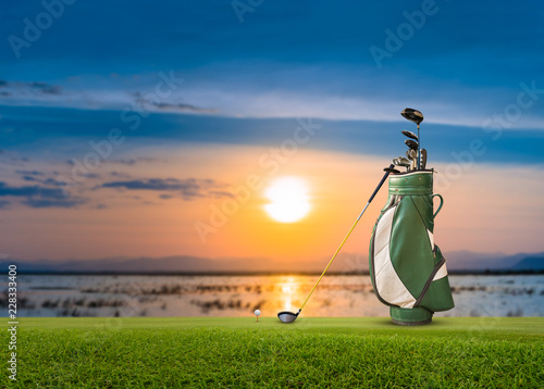 Golf bag, ball, putter and driver (wood no 1) on green sunset as background.