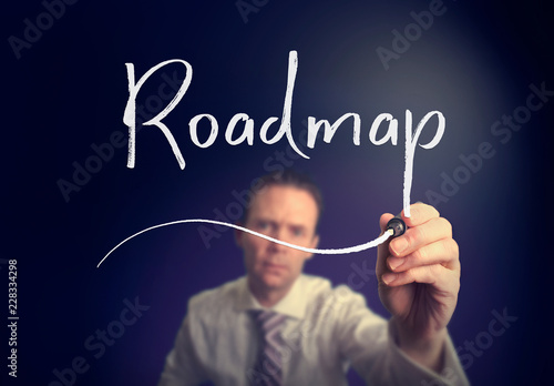 A businessman writing a Roadmap concept with a white pen on a clear screen.