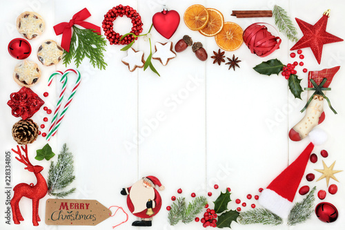 Christmas background border composition with traditional symbols of bauble tree decorations, candy canes, mince pies, fruit, spices, winter flora, ribbon and gift tag on rustic white wood. Top view.