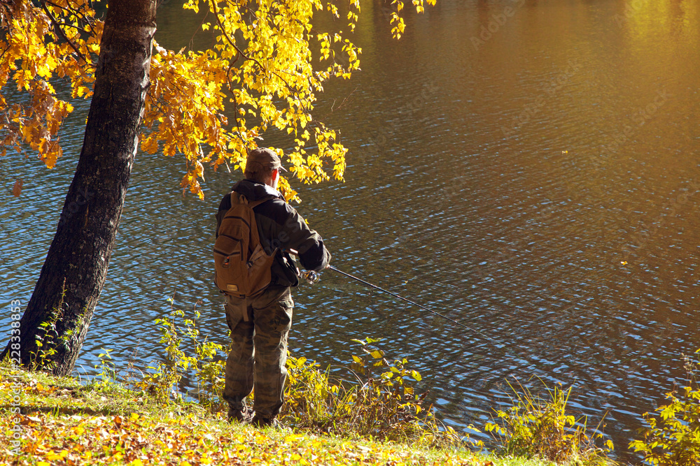 A fisherman with a fishing rod standing on the shore of a lake in sunny autumn weather and birch with yellow leaves A fisherman with a fishing rod standing on the shore of a lake in sunny autumn weather and birch with yellow leaves