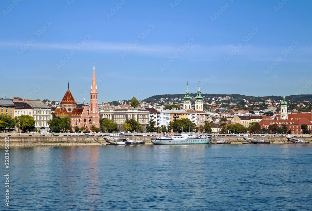 Blue sky water view of Budapest, Hungary and the blue Danube with boats and various buildings along the riverbank.
