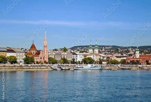 Blue sky water view of Budapest, Hungary and the blue Danube with boats and various buildings along the riverbank.