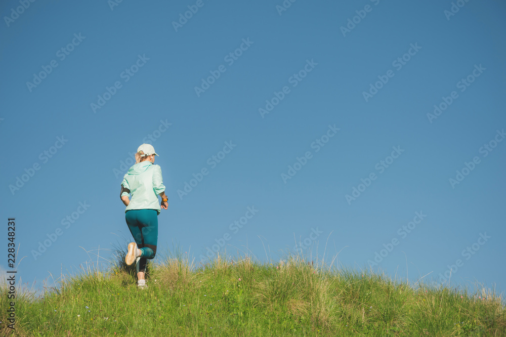 Attractive sports girl in a cap and headphones jogging through the grass in a picturesque place up the hill against the blue sky. Workout outdoors Attractive sports girl in a cap and headphones jogging through the grass in a picturesque place up the hill against the blue sky. Workout outdoors